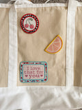 A beige canvas tote bag with white fabric trim and handles, decorated with three embroidered patches: a round cherry patch, a citrus slice patch, and a rectangular patch reading 'I love that for you'.