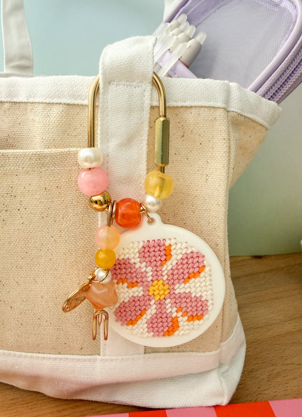 A close-up of a beige canvas tote bag with a decorative cross-stitch flower charm hanging from a gold metal ring. The charm features pink and orange stitched petals with a yellow center, accented by pastel pink, yellow, and white beads, and a small gold bird charm.