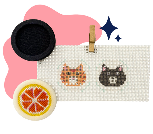 A close-up product shot showing a white cross-stitch sampler with two completed cat face designs, a wooden clothespin, a black circular fabric item, and a round white item with an orange slice pattern, arranged on a white background with pink and blue graphic accents.