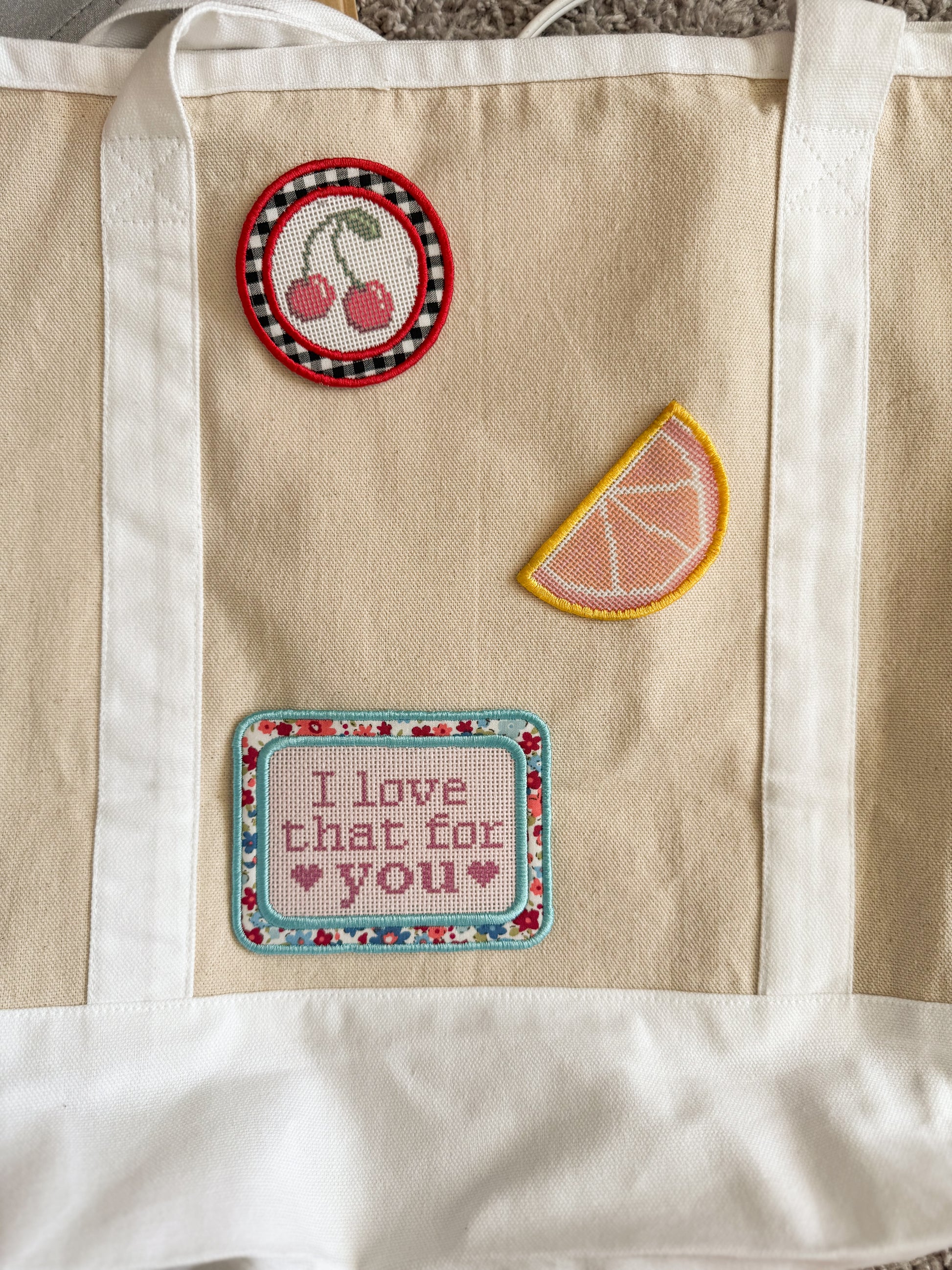 A beige canvas tote bag with white fabric trim and handles, decorated with three embroidered patches: a round cherry patch, a citrus slice patch, and a rectangular patch reading 'I love that for you'.