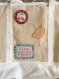 A beige canvas tote bag with white fabric trim and handles, decorated with three embroidered patches: a round cherry patch, a citrus slice patch, and a rectangular patch reading 'I love that for you'.
