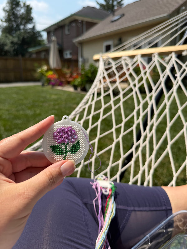 A hand holds a small round embroidered flower ornament with a purple flower and green leaves in front of a backyard scene with a hammock and lawn.