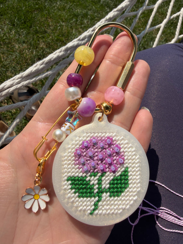 A close-up of a hand holding a decorative embroidered flower keychain or bag charm. The round white embroidered disc features a purple flower with green leaves, attached to gold-toned hardware and colorful beads, with a small white daisy charm dangling below.