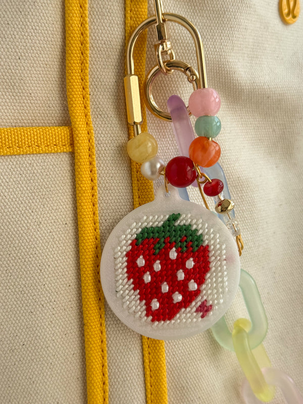A close-up of a cross-stitch style strawberry charm attached to a gold key ring and keychain, featuring colorful beads and translucent pastel pieces on a cream fabric background with yellow stitching.