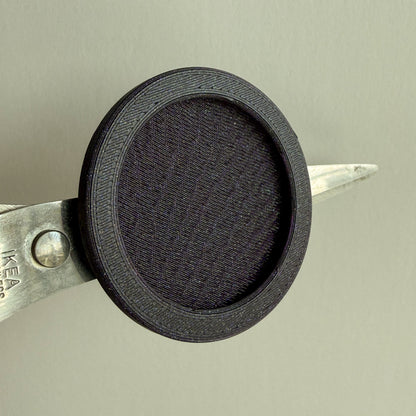 A close-up of a black mesh mini magnet frame attached to the opening of metal scissors against a plain light gray background.