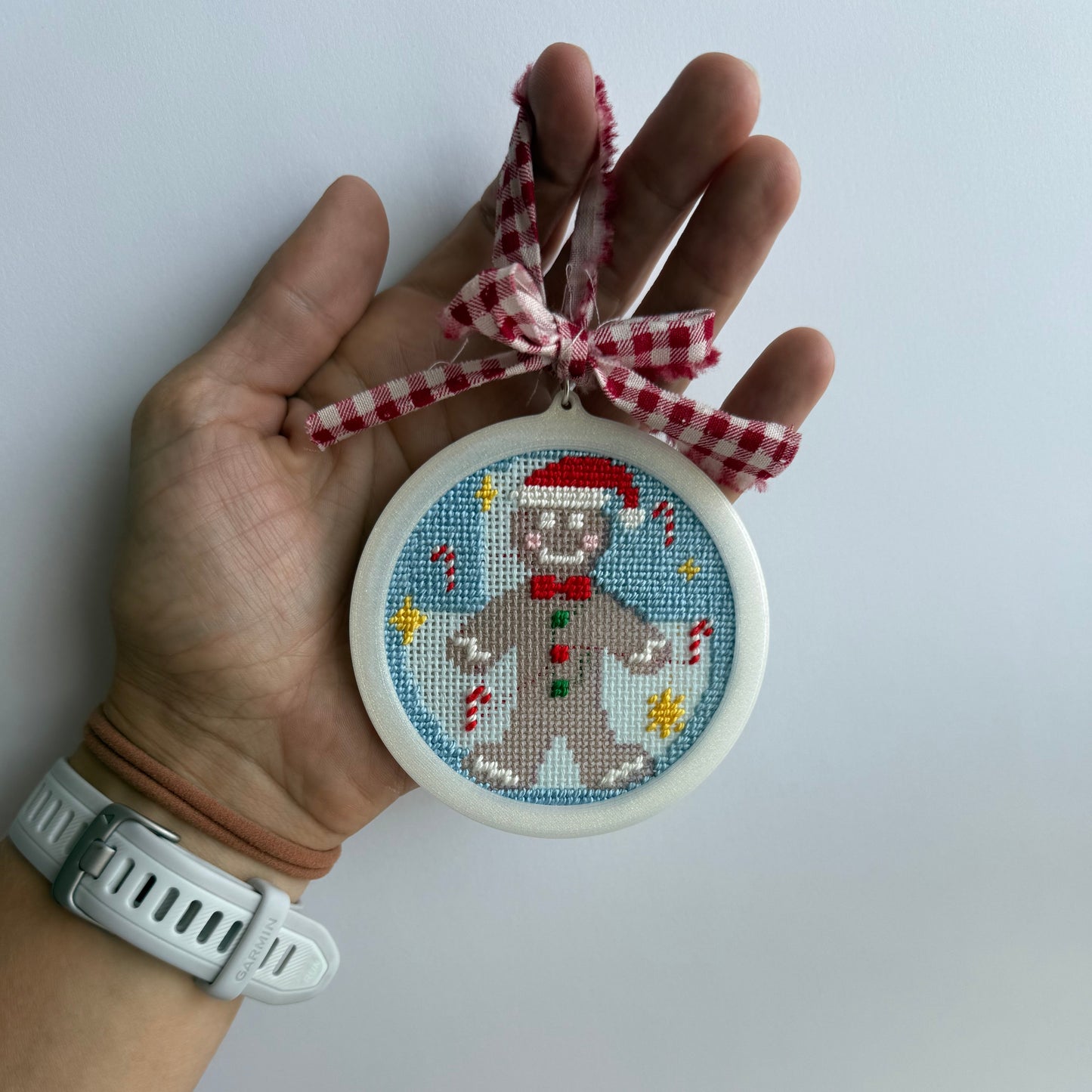 A hand holds a round white ornament featuring a cross-stitched gingerbread man with a red Santa hat and bow tie, hanging from a red-and-white gingham ribbon. The background is a plain light gray wall.
