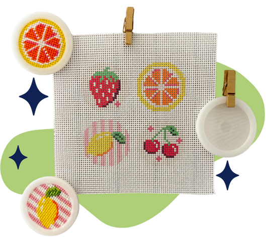 A square white cross-stitch canvas clipped at the top, featuring embroidered fruit motifs including a strawberry, orange slice, cherries, and a lemon. The canvas is displayed on a light green abstract background with decorative blue star shapes and three round white framed pieces around it.