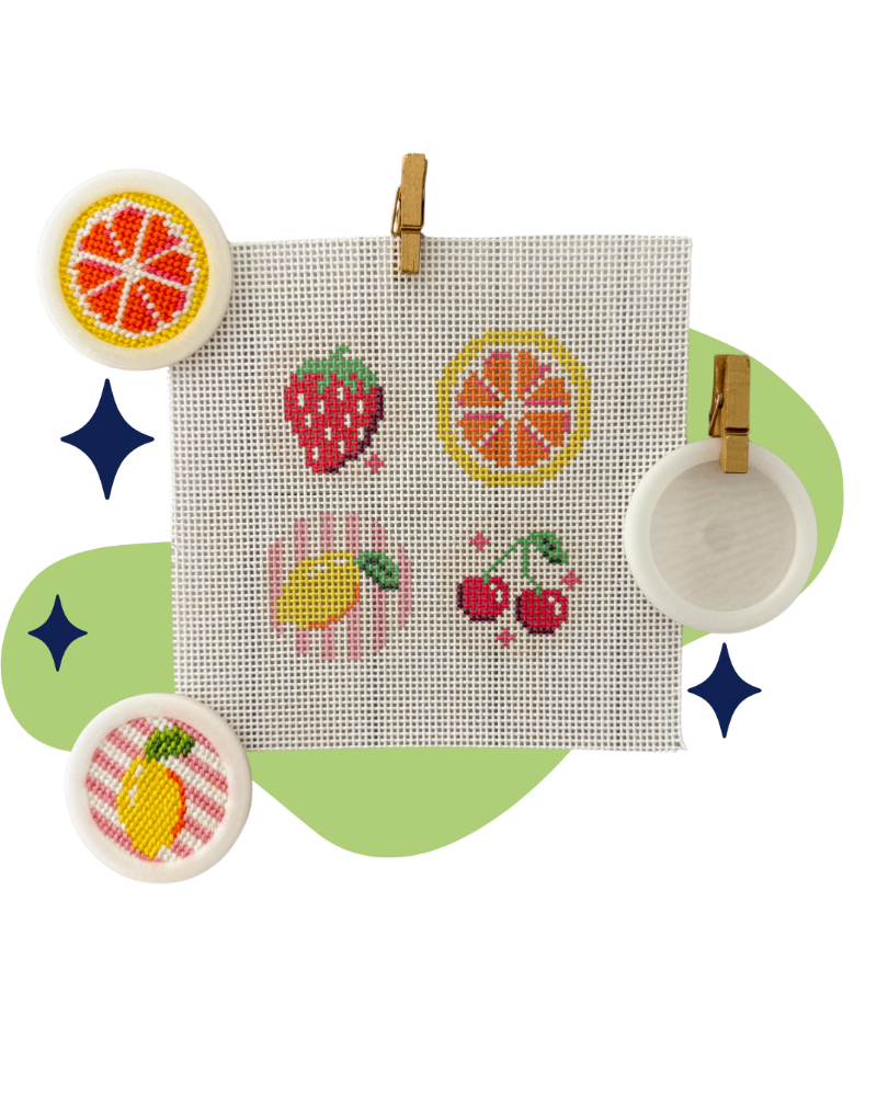 A square white cross-stitch canvas clipped at the top, featuring embroidered fruit motifs including a strawberry, orange slice, cherries, and a lemon. The canvas is displayed on a light green abstract background with decorative blue star shapes and three round white framed pieces around it.